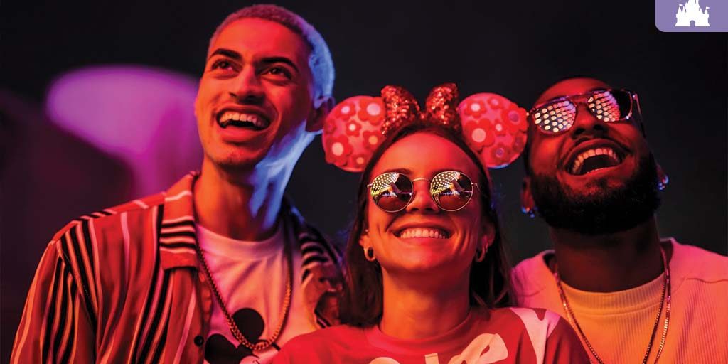 Three friends wearing Disney-themed outfits and sunglasses smile under colorful nighttime lights at a Disney After Hours event.