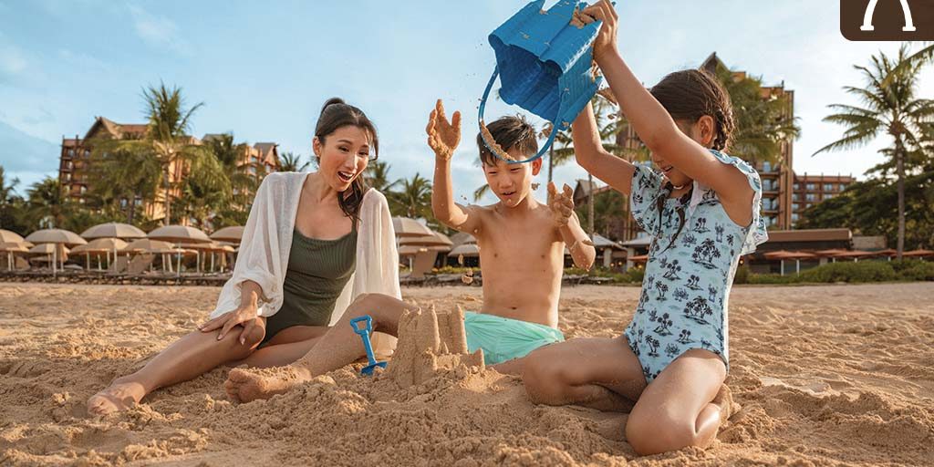 Family building a sandcastle on the beach at Aulani, A Disney Resort & Spa, with palm trees and resort buildings in the background.