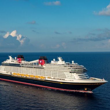 disney-cruise-line-wish-at-sea-2 A large cruise ship sails through calm blue waters under a clear sky with fluffy clouds in the background.