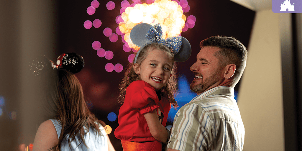 Smiling father holding daughter in Mickey ears while watching colorful fireworks at Walt Disney World Resort.