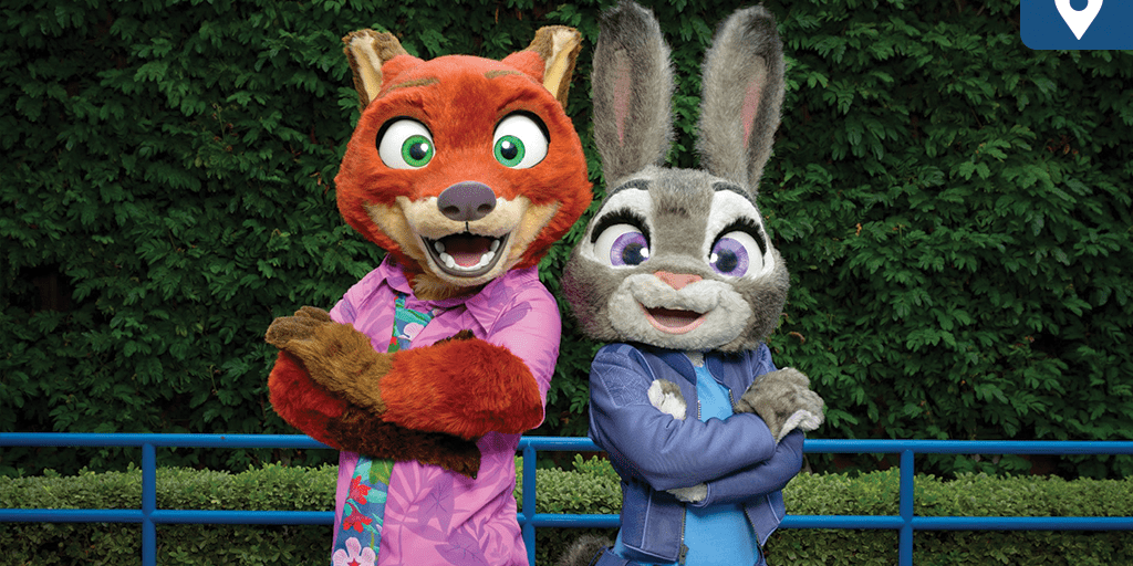 Nick Wilde and Judy Hopps pose back-to-back in their Zootopia 2 outfits at a Disney park, smiling in front of lush greenery.