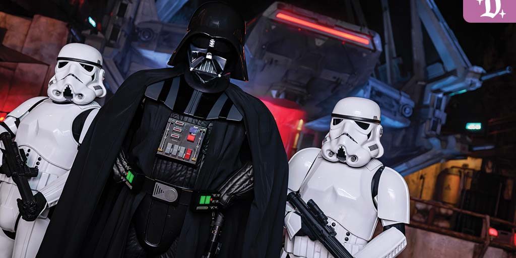 Darth Vader stands flanked by two Stormtroopers at Galaxy’s Edge, with a TIE fighter and Batuu backdrop illuminated at night.