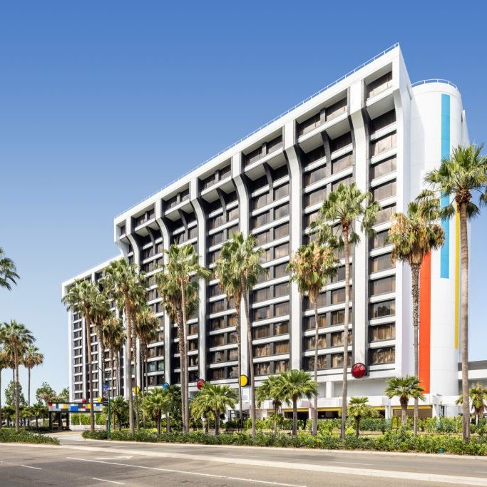 A modern hotel building with a colorful facade, surrounded by palm trees, set against a clear blue sky.