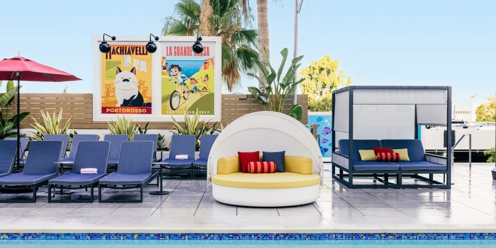 A vibrant poolside scene featuring blue loungers, a round white daybed with colorful pillows, and palm trees against a clear sky.