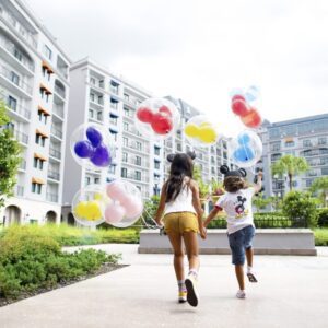 Two children run through a park, joyfully carrying colorful balloons, with modern buildings and greenery in the background.