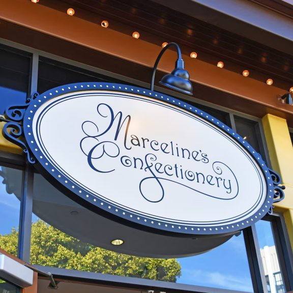 Sign hanging above a storefront reads "Marceline's Confectionery," with decorative elements and warm lighting outside.