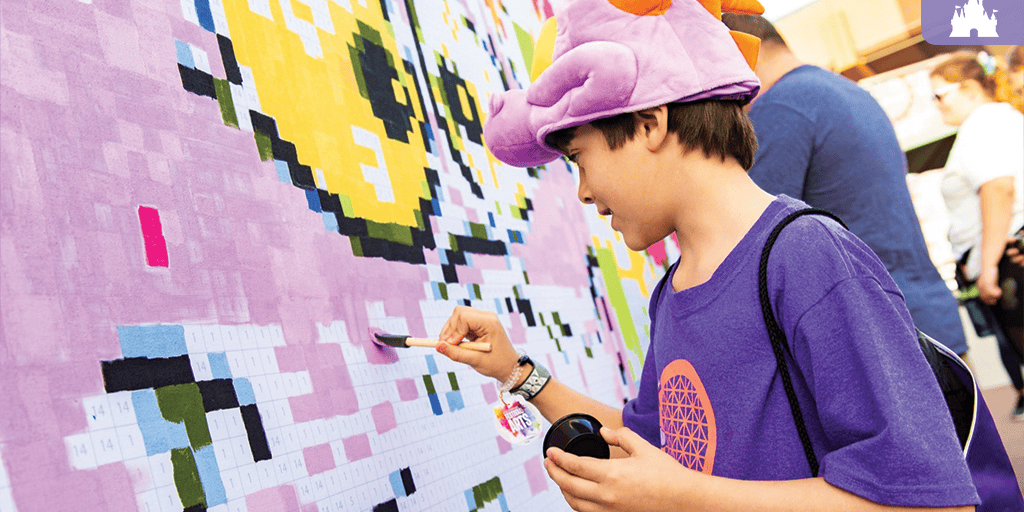 Child in purple shirt and dragon hat paints numbered squares on a large outdoor pixel-art mural with a brush and paint palette.