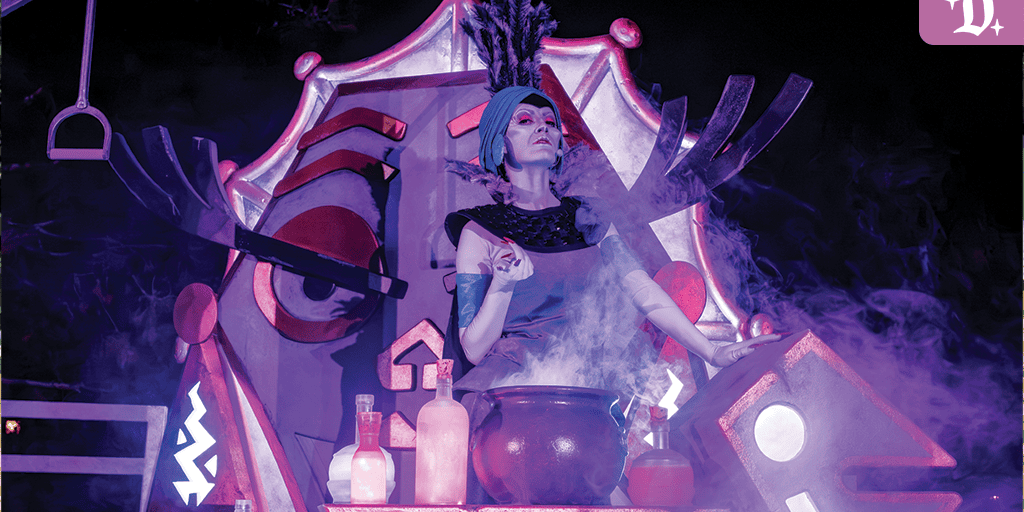 Performer in theatrical costume stands behind a smoking cauldron on stage decorated with a large stylized mask and glowing potion bottles