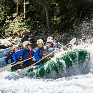 A group of people in blue jackets and helmets paddles a green raft through a whitewater river, surrounded by trees.