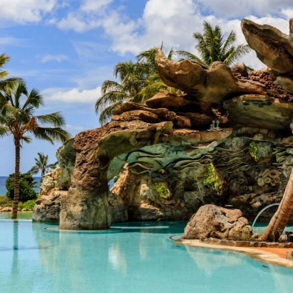 A tranquil pool area surrounded by palm trees and rocky formations, with ocean views under a blue sky with fluffy clouds.