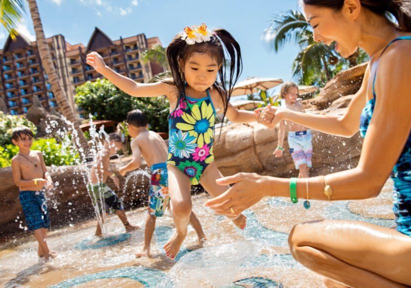 A girl in a colorful swimsuit jumps into a splash pad while a woman reaches out to catch her, with kids playing and a resort in the background.