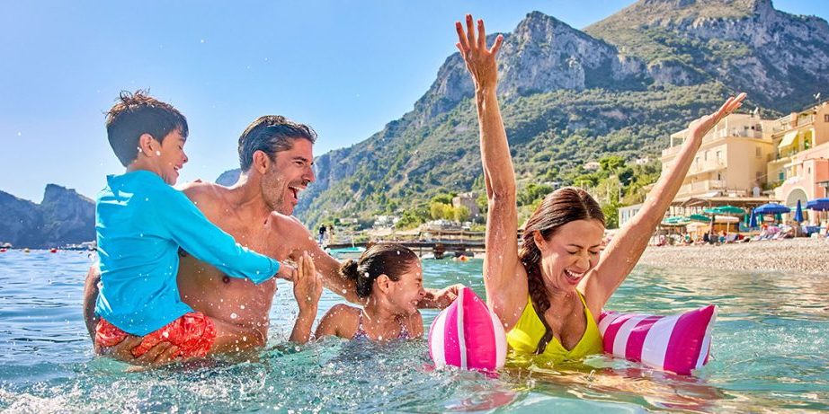 Family playing in ocean waters with mountain coastal town in background, celebrating a summer cruise vacation