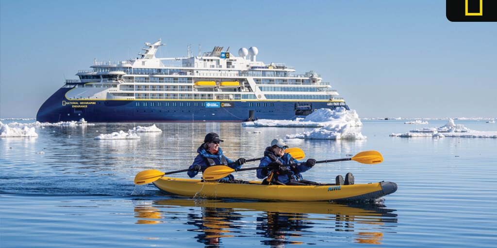 nat-geo-expeditions-2027-2028 Two people kayak through icy Arctic waters with the National Geographic Endurance expedition ship in the background under clear blue skies.