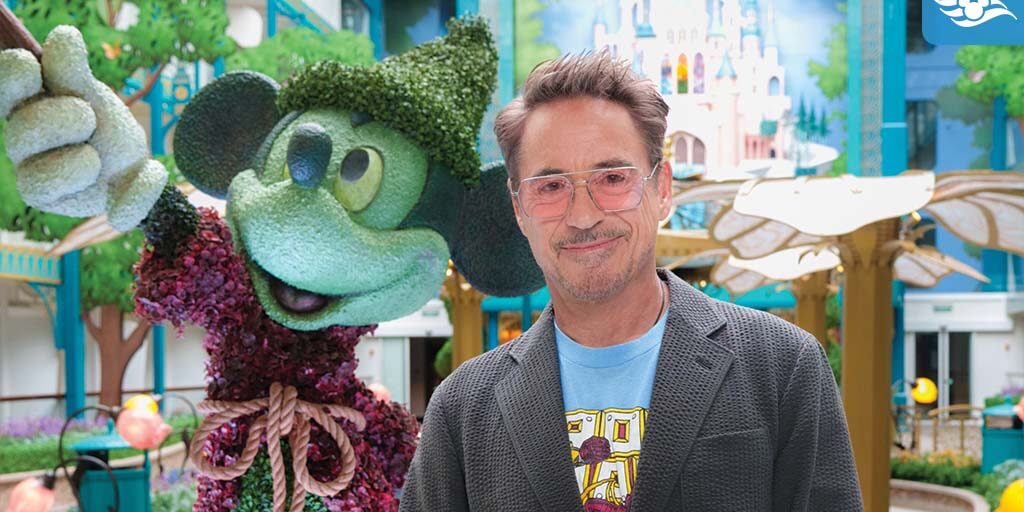 Robert Downey Jr. poses beside a Mickey Mouse topiary aboard Disney Adventure, with colorful Disney castle artwork in the background.