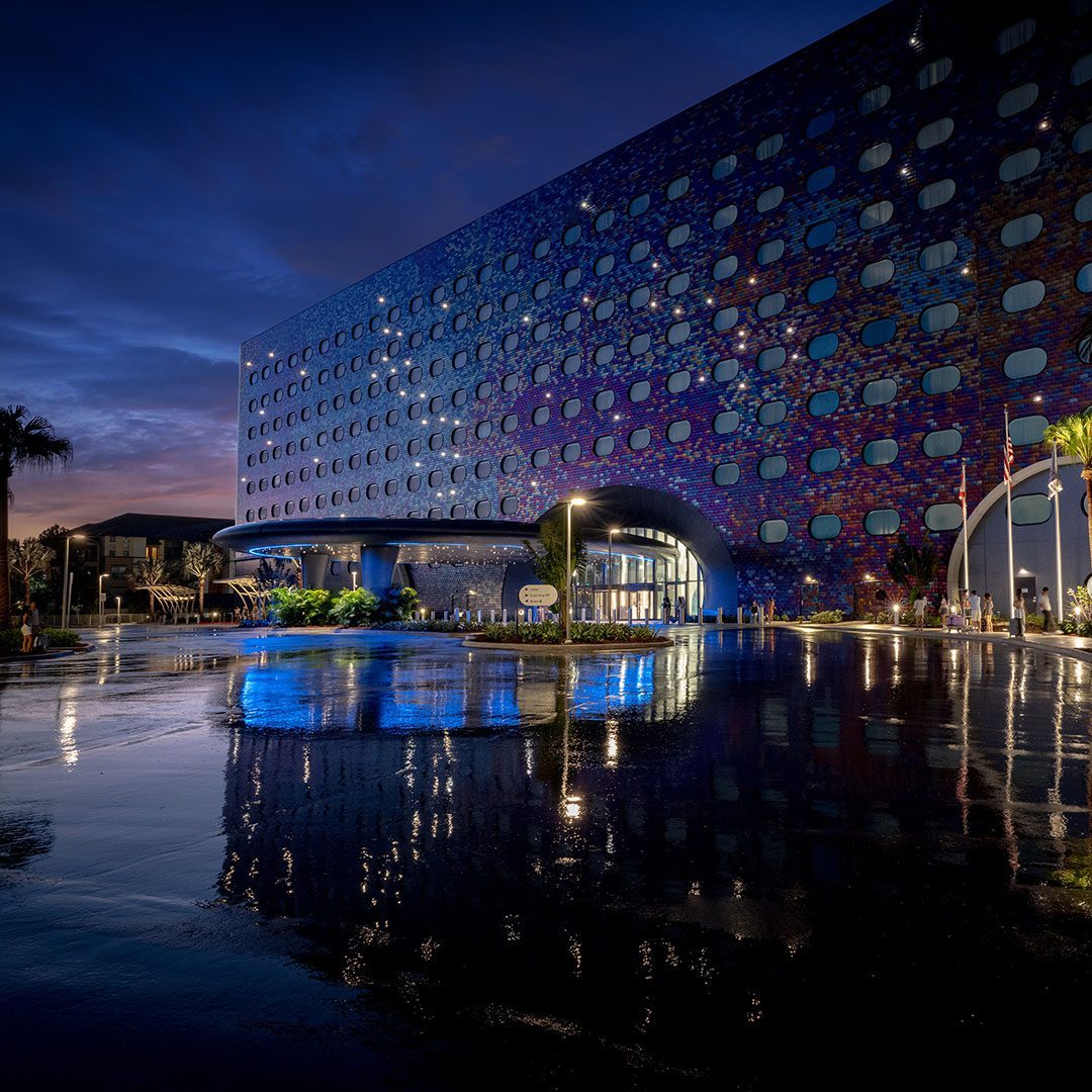 Tall nighttime view of a futuristic hotel, showcasing its shimmering facade and circular entrance lit against a dark sky.
