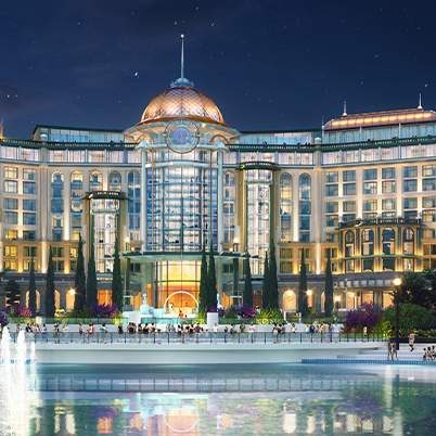 A grand hotel illuminated at night, featuring ornate architecture and a fountain in front, surrounded by lush greenery.