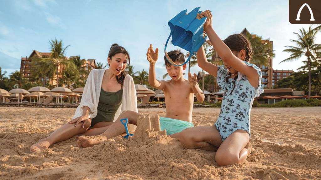 Family building a sandcastle on the beach at Aulani, A Disney Resort & Spa, with palm trees and resort buildings in the background.