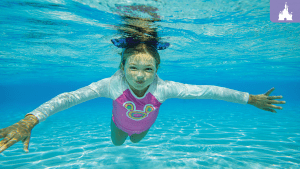 Young girl swimming underwater at Disney's Typhoon Lagoon, wearing a pink rash guard with a Mickey design and a bow headband.