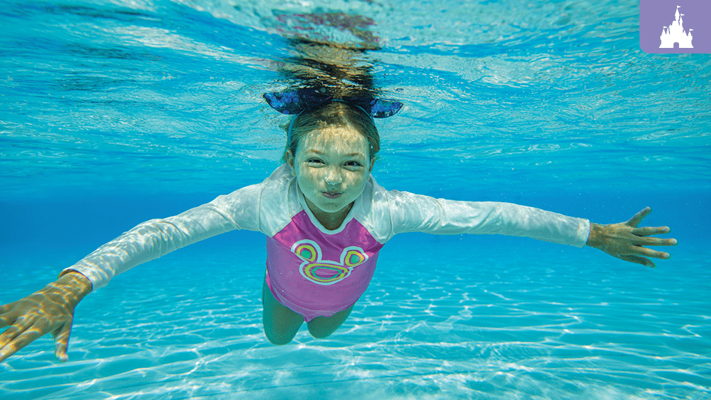 disney-blizzard-beach-typhoon-lagoon Young girl swimming underwater at Disney's Typhoon Lagoon, wearing a pink rash guard with a Mickey design and a bow headband.