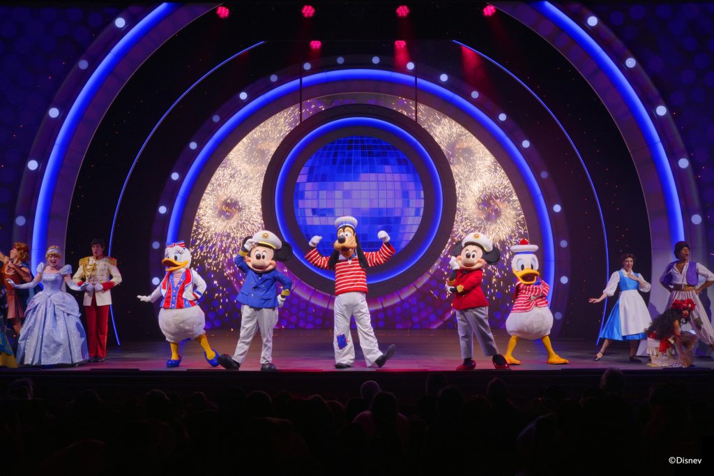 Goofy leads a vibrant stage performance in Disney Cruise Line's "Seas the Adventure" show, surrounded by Disney characters in colorful costumes.