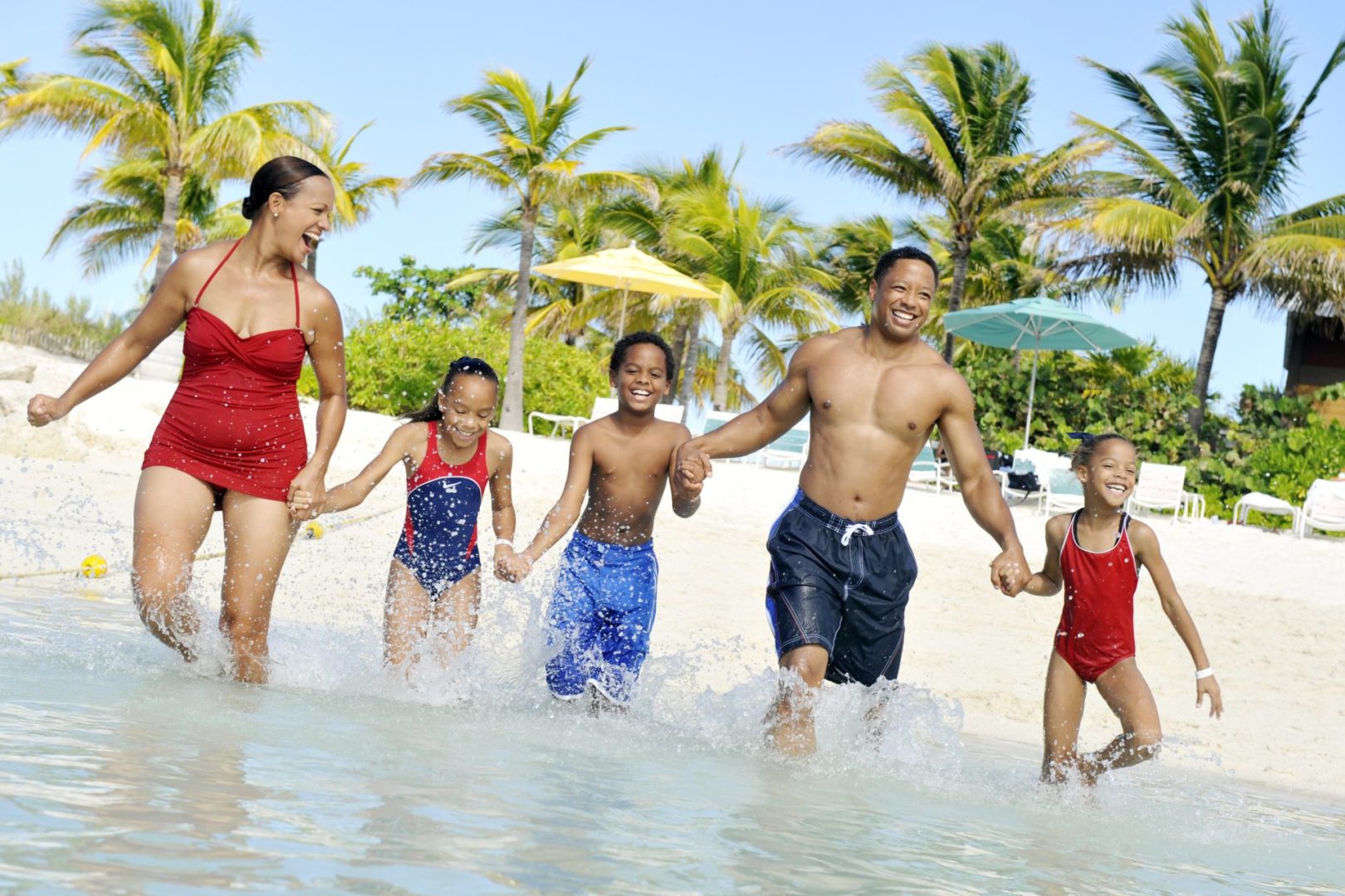 A joyful family of five plays together in shallow water at the beach, surrounded by palm trees and colorful umbrellas.