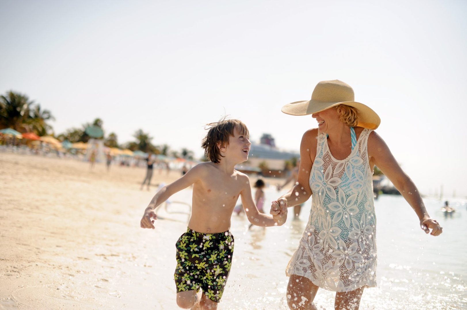 A woman and a young boy run joyfully along the beach, splashing in the water under a sunny sky.
