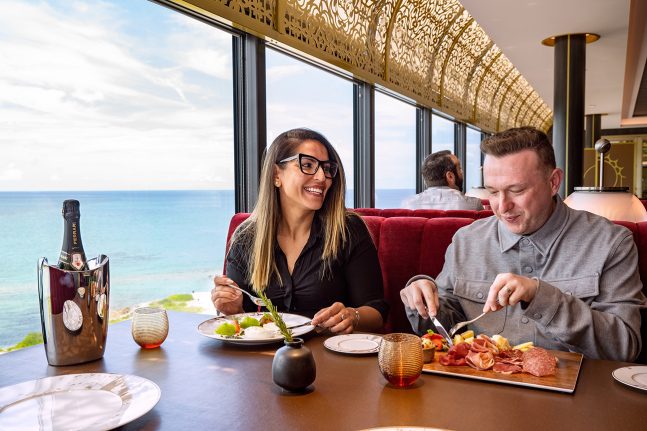 A man and woman are dining together at a table with a scenic ocean view, enjoying gourmet food and drinks, with a stylish interior.