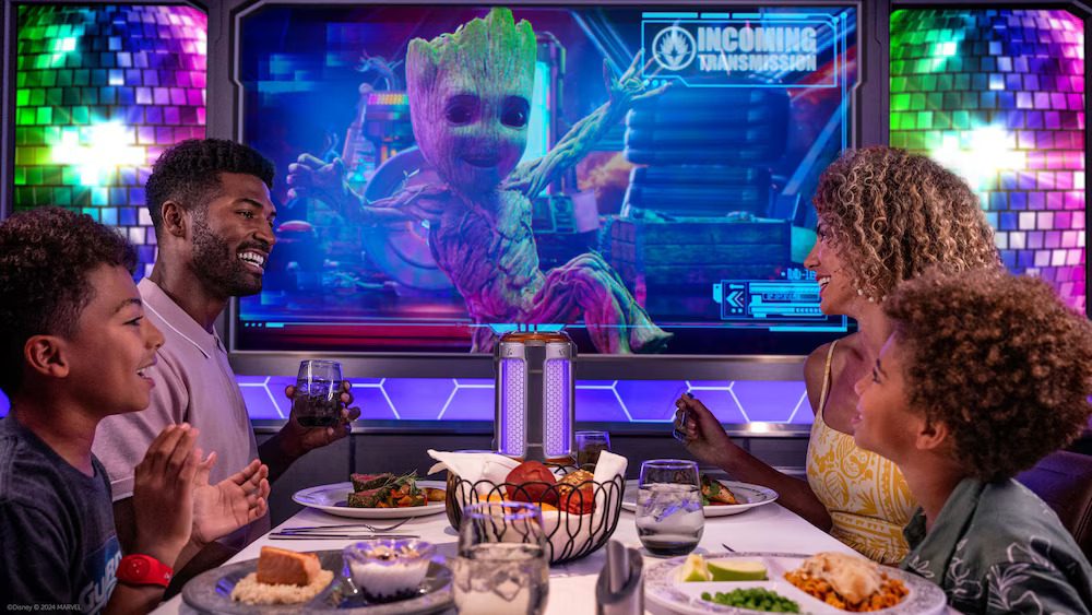 A family enjoys a meal while watching a colorful screen featuring a character resembling Baby Groot, surrounded by vibrant decorations.