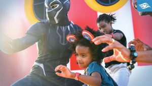 A child in a green shirt excitedly interacts with a costumed superhero, while another child and an adult join in the fun.