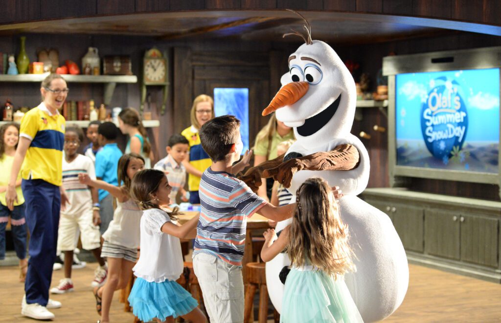A large snowman character stands playfully with children around him in a lively indoor setting, with adults and a TV in the background.