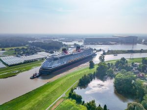 Aerial view of the Disney Treasure cruise ship being guided by a tugboat down the narrow Ems River, surrounded by lush green landscapes and industrial buildings in the background.