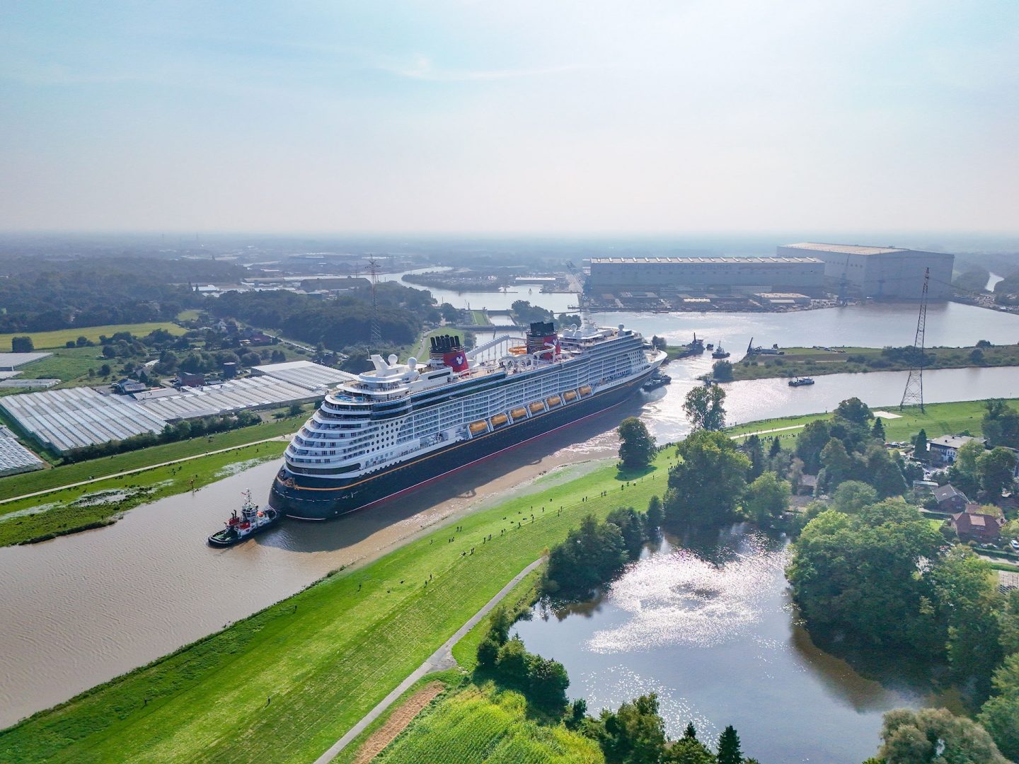 disney-cruise-line-treasure-conveyance Aerial view of the Disney Treasure cruise ship being guided by a tugboat down the narrow Ems River, surrounded by lush green landscapes and industrial buildings in the background.