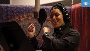 A smiling person wearing a black outfit and headset sings passionately in a recording studio, with a colorful backdrop.