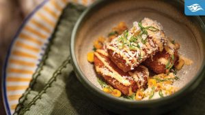 A bowl of golden-brown fritters topped with cheese, herbs, and corn, served on a patterned plate with a green linen napkin.