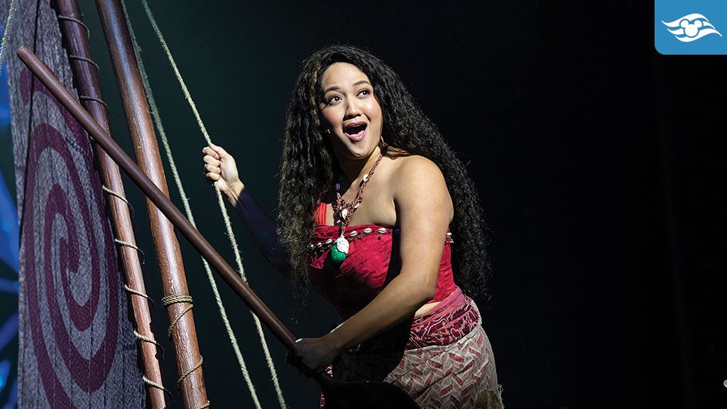 Moana performing on stage during Disney The Tale of Moana aboard the Disney Treasure, standing on a canoe with a sail adorned with a red spiral design, exuding determination and joy.