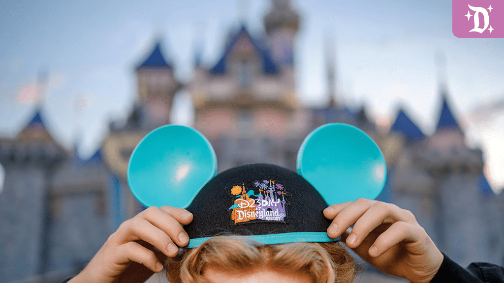 disney-d23-2024 A person is holding a blue hat with mouse ears in front of a castle, ready to put it on at a theme park.
