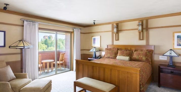 Cozy hotel room featuring a wooden bed, armchair, and lamps, with large windows opening to a balcony and scenic views.