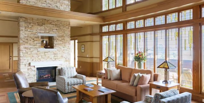 A cozy living room with large windows, stone fireplace, and comfortable furniture, featuring warm wood accents and decorative lamps.