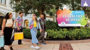A group of friends walking and smiling at Disney Springs, carrying shopping bags, near a colorful sign that reads "Be Healthy, Be Well" for Wellness Month presented by AdventHealth.
