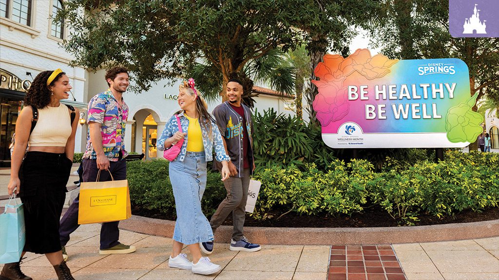 disney-springs-wellness-journey A group of friends walking and smiling at Disney Springs, carrying shopping bags, near a colorful sign that reads "Be Healthy, Be Well" for Wellness Month presented by AdventHealth.