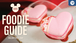 A close-up of pink heart-shaped macarons decorated with sprinkles, accompanied by text: "Foodie Guide" for Valentine's Day. Mickey Mouse logo included.