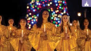 Choir members in golden robes holding candles in front of a brightly lit Christmas tree at EPCOT’s Candlelight Processional.