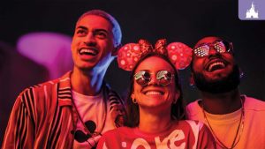 Three friends wearing Disney-themed outfits and sunglasses smile and enjoy vibrant nighttime lights at a Disney After Hours event.