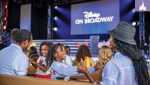 A family seated on wooden benches smiles and holds hands while enjoying a Disney on Broadway performance at EPCOT, with the stage and logo visible in the background.