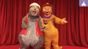 Two colorful, anthropomorphic animal characters stand on stage, singing joyfully in front of a red curtain backdrop.