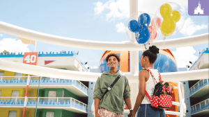 Smiling couple holding Mickey Mouse balloons at Disney’s All-Star Sports Resort, surrounded by colorful buildings and whimsical decor.