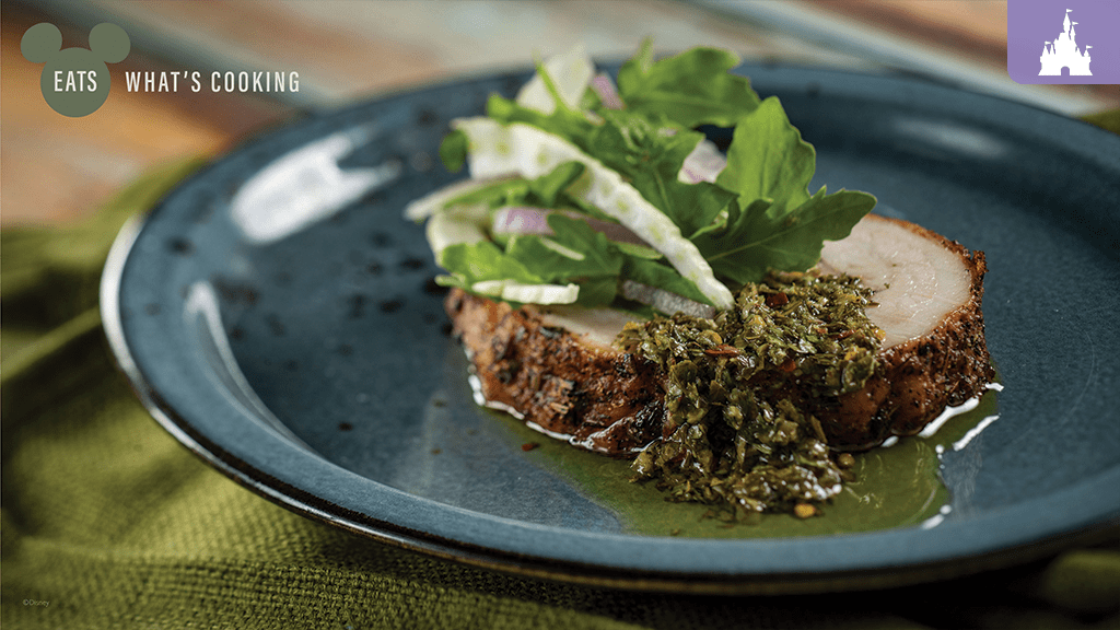 disney-world-epcot-festival-marketplaces-menu A slice of seasoned meat topped with green herb sauce and fresh salad, served on a blue plate with a textured green backdrop.