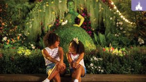 Two young girls sit on a bench, smiling at each other, surrounded by lush greenery and colorful flowers. Behind them, a topiary of Princess Tiana from The Princess and the Frog glows under string lights, creating a magical nighttime atmosphere.