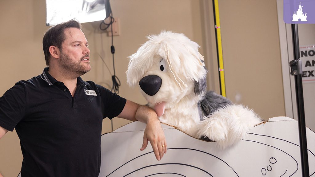 A man in a black shirt leans beside a large, fluffy dog puppet on a set, engaged in conversation against a studio backdrop.