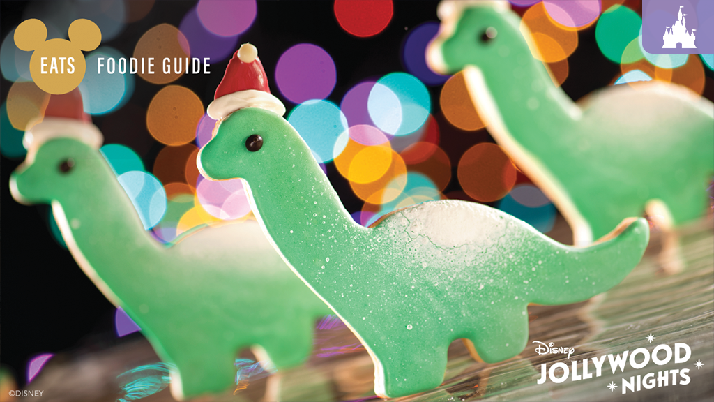 Three green dinosaur cookies wearing Santa hats against a colorful, blurred background with holiday lights. Text overlay includes "EATS" and "JOLLYWOOD NIGHTS."